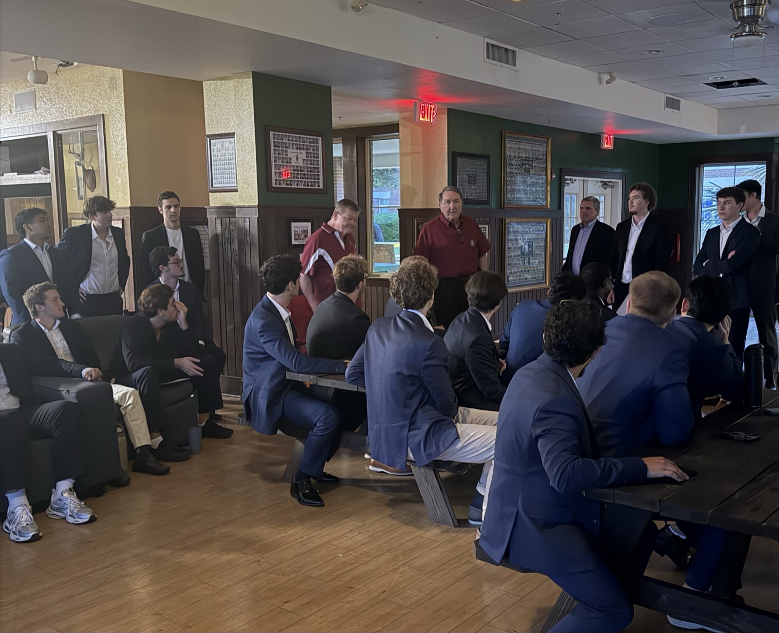 Brothers in jackets seated and standing around the chapter room, listening to alumni speaking at the front